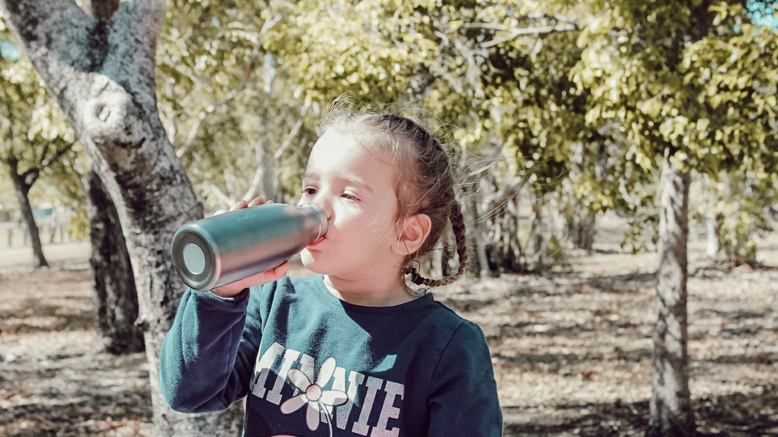 Ein kleines Mädchen trinkt aus einer 720°DGREE milkyBottle, im Hintergrund sind einige Bäume zu sehen, die von der Sonne angestrahlt werden. Aus allen 720°DGREE Flaschen ist das Trinken ein Kinderspiel und macht sogar Spaß - Ideal für Kindergarten Kinder.